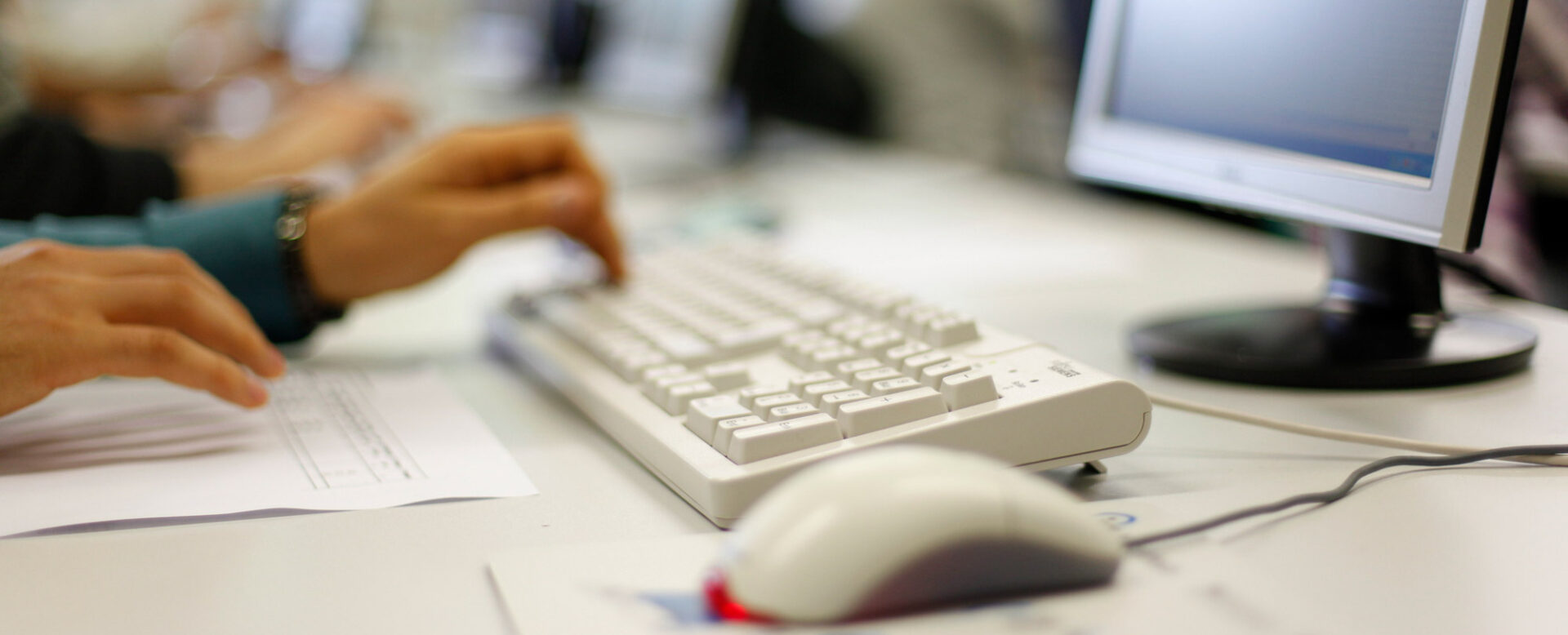 Person tippt auf Tastatur vor einem Computerbildschirm in einem Büro oder Klassenzimmer.
