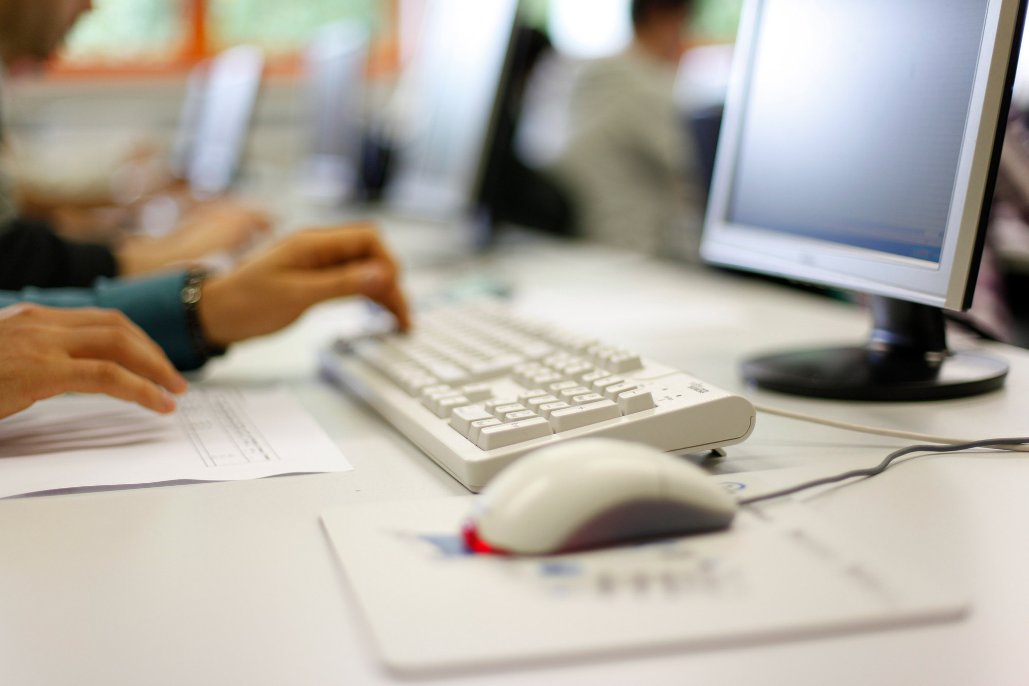 Person tippt auf Tastatur vor einem Computerbildschirm in einem Büro oder Klassenzimmer.