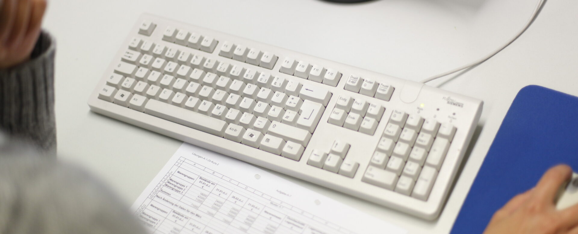 Person arbeitet an einem Computer mit einer weißen Tastatur und Maus auf einem blauen Mauspad. Ein Dokument liegt auf dem Tisch.