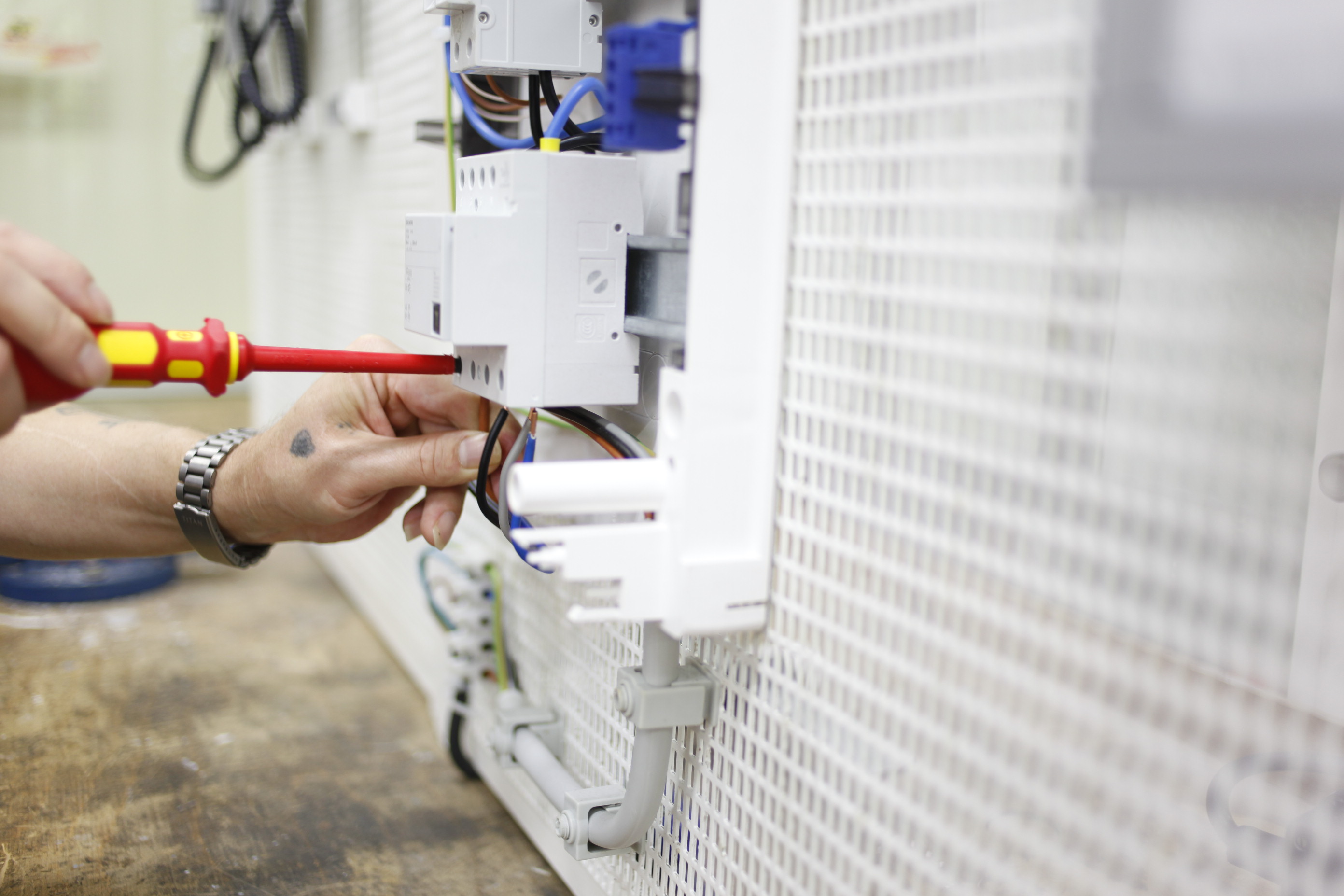 Person arbeitet mit Schraubenzieher an elektrischer Schalttafel, Kabel sichtbar. Hände fokussiert, Detailarbeit.