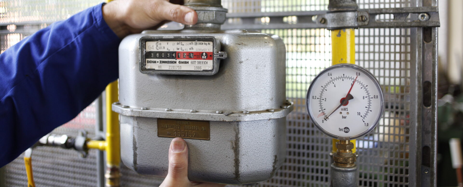 Hände kontrollieren einen Gaszähler mit einer sichtbaren Druckanzeige an einem gelben Rohr in einer technischen Umgebung.
