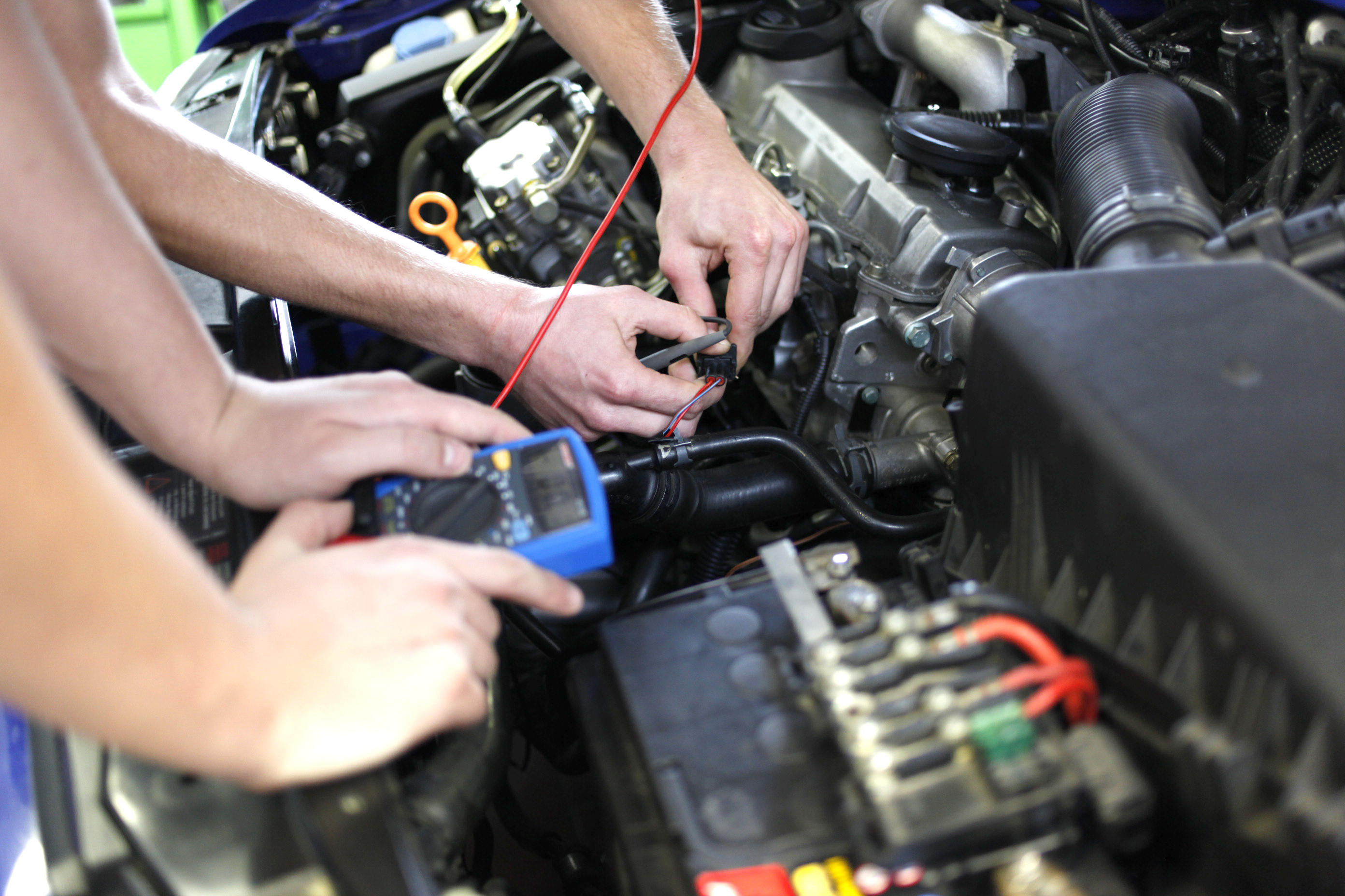 Zwei Personen prüfen mit einem Multimeter die Kabel einer Autobatterie im Motorraum.