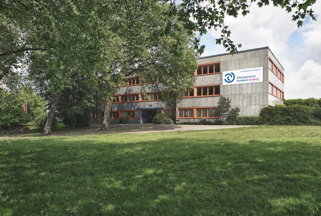 Großes Gebäude mit rotem Fensterrahmen im Grünen, Schild Bildungszentrum Handwerk Duisburg an der Fassade.
