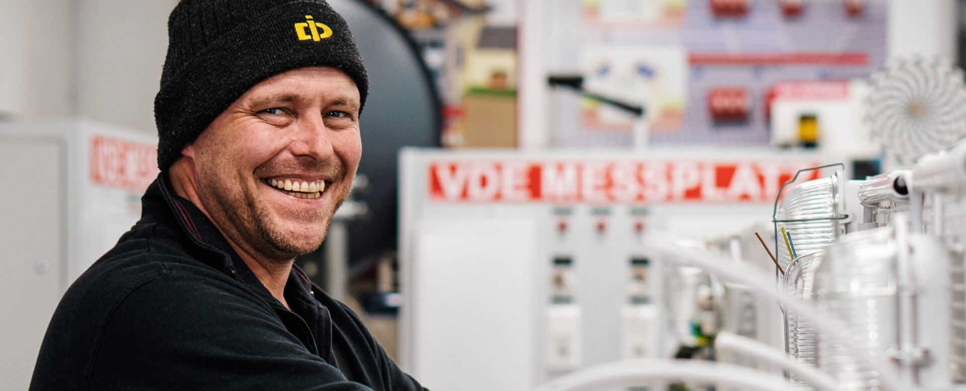 Ein lachender Mann in Arbeitskleidung arbeitet an elektrischen Schaltanlagen in einer Werkstatt.