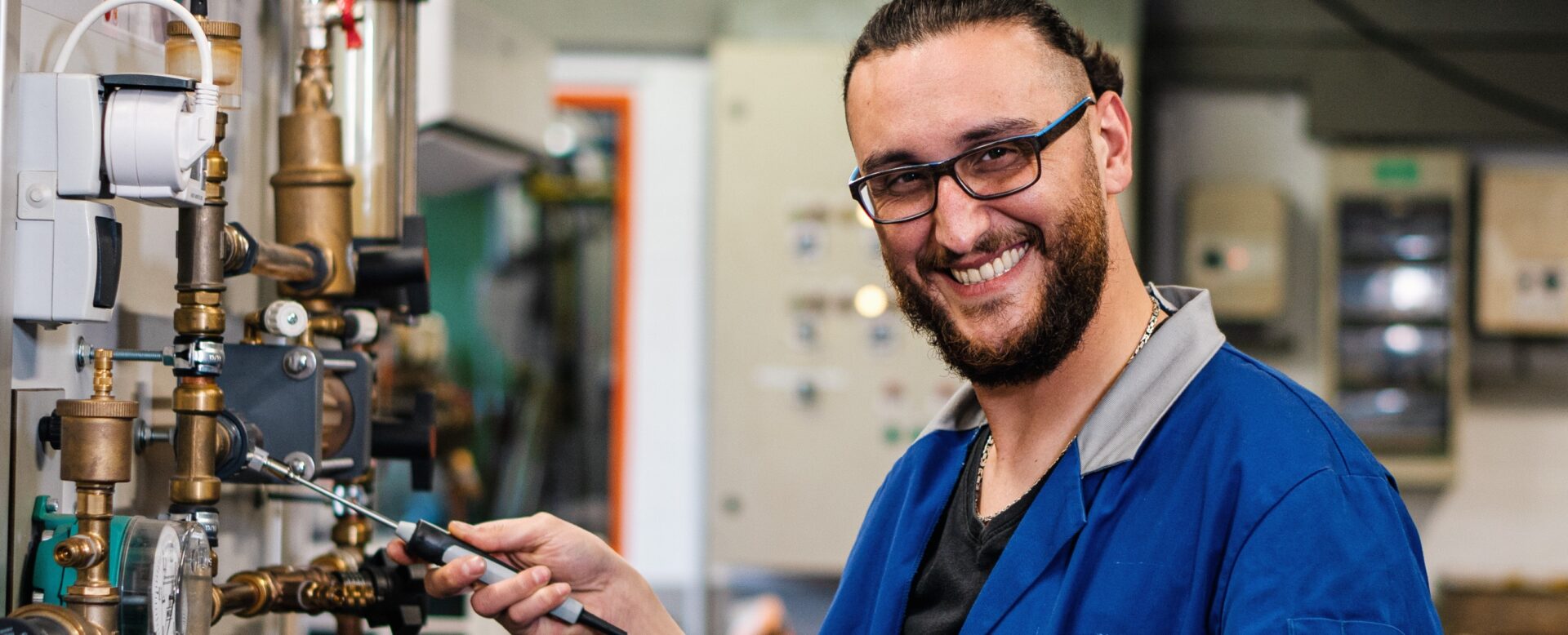 Ein Techniker im blauen Kittel arbeitet lächelnd an einem System von Rohren und Ventilen, hält Messgeräte in der Hand.