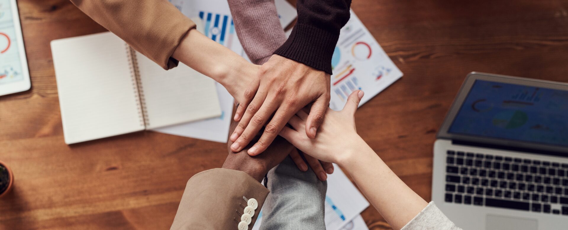 Fünf Personen legen ihre Hände in der Mitte auf einem Tisch, um Zusammenarbeit und Teamarbeit zu symbolisieren.